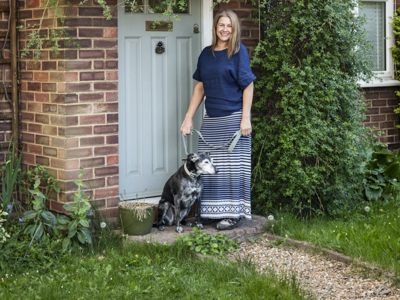 A dog sitter and a dog stand in front of a house with lots of greenery and a garden path