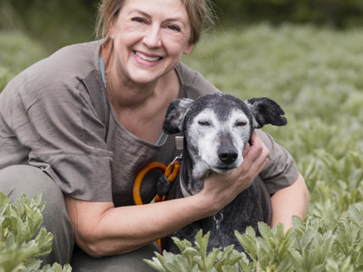 The founder of Mutt and Manor crouches down next to a whippet dog in a green field
