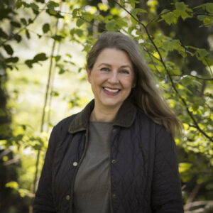 The founder of Mutt and Manor pet sitting service in Holmes Chapel with long brown hair wearing a brown quilted jacket smiles with a woodland scene in the background